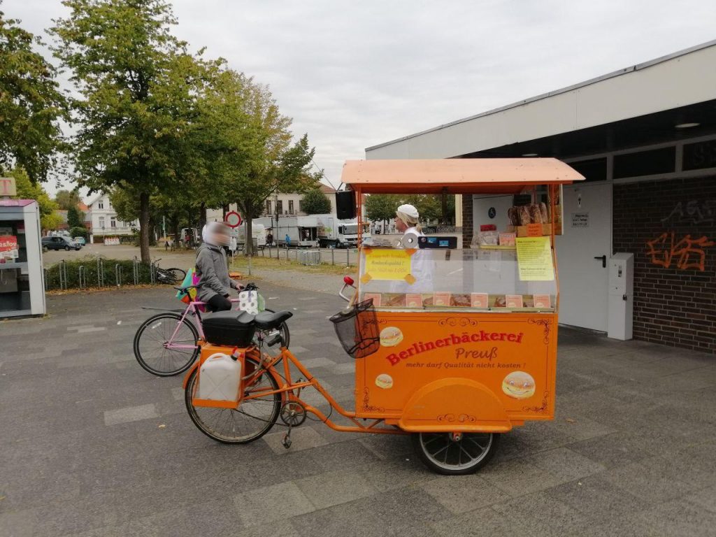 Ein orangenes Lastenfahrrad, mit der Aufschrift "Berlinerbäckerei Preuß" steht auf dem Platz vor dem Kiosk am Pferdemarkt