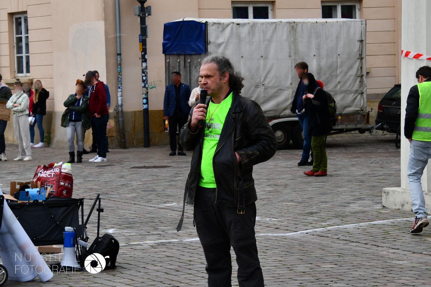 Person mit Mikrofon auf dem Schlossplatz, im Hintergrund mehrere Passant*innen